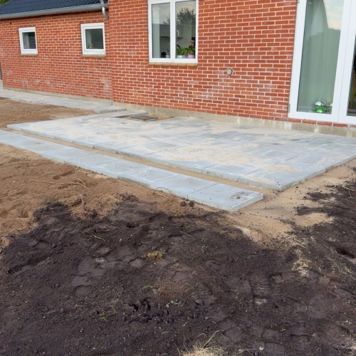 Flisearbejde med ny terrasse foran rødt murstenshus og mørkt tegltag i Tjæreborg.