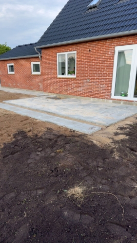 Flisearbejde med ny terrasse foran rødt murstenshus og mørkt tegltag i Tjæreborg.